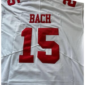 May include: White jersey with red lettering and numbers. The name "BACH" is displayed above the number "15" on the front. The number "5" is visible on the shoulders. The jersey is made of a smooth, likely synthetic, fabric.