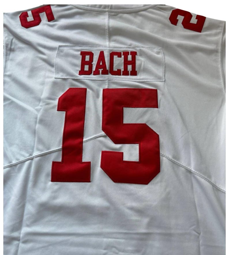 May include: White jersey with red lettering and numbers. The name "BACH" is displayed above the number "15" on the front. The number "5" is visible on the shoulders. The jersey is made of a smooth, likely synthetic, fabric.