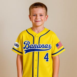 Op de afbeelding: Geel honkbalshirt met blauwe bies en het woord "Bananas" in blauw schrift. Het shirt heeft het nummer "4" op de voorkant en een Amerikaanse vlag op de mouw. Op de andere mouw staat een "LOGO" patch.