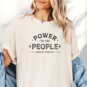 以下が含まれることがあります： 「POWER - TO THE - PEOPLE」の文字がダークなヴィンテージフォントでプリントされたクリーム色のTシャツ。 「SHOW UP SPEAK OUT」というフレーズも含まれています。 Tシャツはデニムジャケットと組み合わせています。