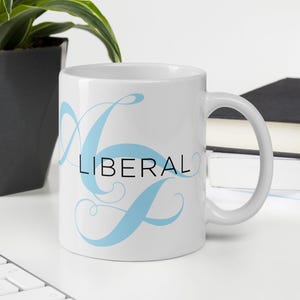 May include: White ceramic mug with the word "LIBERAL" in black text and light blue decorative script. The mug is on a white surface with a plant and books in the background.