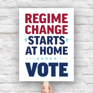 May include: A white sign with the words "REGIME CHANGE STARTS AT HOME VOTE" in red and blue lettering. The sign is held by a wooden stick against a white brick wall.