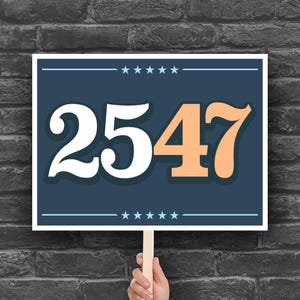 May include: A rectangular sign displaying the number "2547" in white and orange against a dark blue backdrop. The sign is framed in white and held by a wooden stick. The background is a dark grey brick wall.