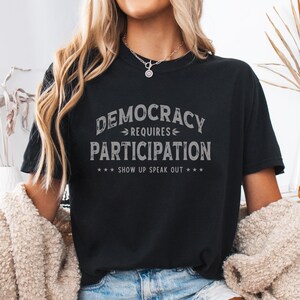 以下が含まれることがあります： グレーの文字で「DEMOCRACY REQUIRES PARTICIPATION」と書かれた黒いTシャツ。Tシャツには、星のアクセントが付いた「SHOW UP SPEAK OUT」というフレーズも含まれています。Tシャツはカジュアルなスタイルです。