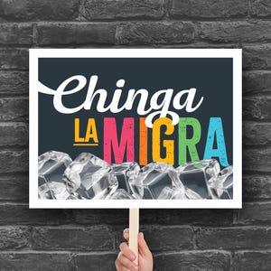 May include: A white sign with the words "Chinga La Migra" in white and colorful letters, with ice cubes at the bottom. The sign is held by a person in front of a brick wall.