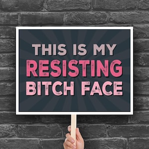 May include: A sign held by a person with the text "THIS IS MY RESISTING BITCH FACE" in pink and white letters on a dark grey background with radiating lines. The sign is on a wooden stick, against a brick wall.