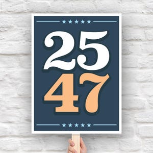 May include: A rectangular sign featuring the numbers "25" in white and "47" in peach against a navy blue backdrop. The sign has a white border and is held by a wooden stick. The top and bottom of the sign have a line of stars.