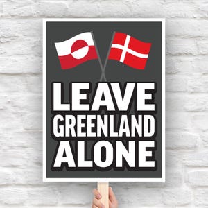 May include: A sign with the text "LEAVE GREENLAND ALONE" in bold white letters on a black background. The sign features crossed flags of Greenland and Denmark at the top, held by a wooden stick against a white brick wall.