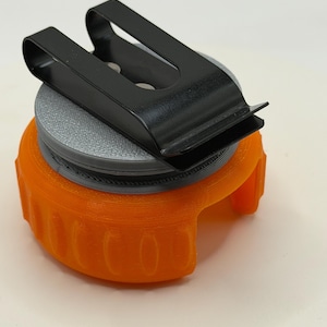 May include: A bright orange circular plastic holder with a textured surface and a vertical opening, topped with a flat gray circular disc. A black metal U-shaped belt clip is securely attached to the top of the gray disc, featuring a smooth finish.