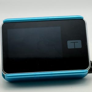 May include: A rectangular medical device with a black screen and a light blue frame. The device has a small white "T" symbol in the lower right corner of the screen. A thin, clear tube extends from the side.
