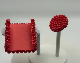 May include: A red decorative object with a square shape and a beaded border, and a red sphere on a white stick. The square object has diagonal lines. The objects are on a white base.