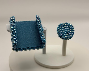 May include: A teal-colored phone stand and a matching pen. The phone stand has a teal fabric base and is framed with iridescent teal pearls. The pen has a round, pearl-covered top. Both items are displayed on white stands.