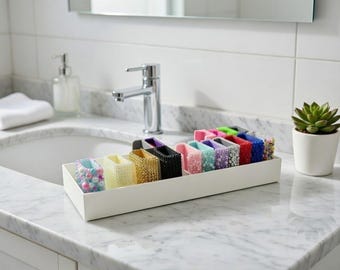 May include: A white rectangular tray filled with colorful, decorative watch band cases. The cases are in various colors, including pink, blue, green, and red, and are embellished with beads and other decorative elements. The tray sits on a marble countertop.