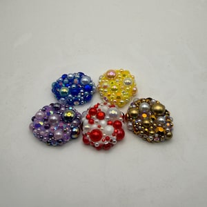 May include: Five circular decorative cabochons covered in a dense arrangement of small beads, pearls, and faceted crystals. The set includes one blue, one yellow, one purple, one red and white, and one bronze and gold-toned cluster piece.