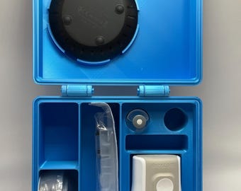 May include: Open blue case with compartments containing medical devices. The case holds a black circular device, a white and gray device with a button, a roll of tape, and other medical supplies. The case is open, revealing the contents.