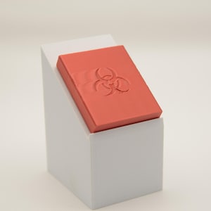 May include: A rectangular, coral-colored object with a biohazard symbol is displayed on a white, angled stand. The stand is a simple, geometric shape, and the background is plain white.