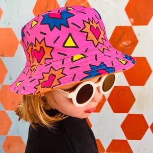 Könnte beinhalten: Ein rosa Bucket-Hut mit einem bunten geometrischen Muster mit Herzen, Sternen und Dreiecken. Der Hut hat einen weißen Rand und ist aus einem leichten Stoff gefertigt.