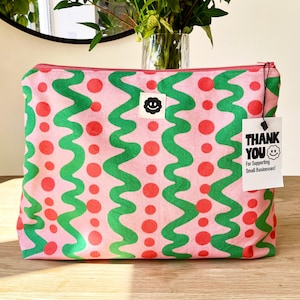 Pode incluir: Bolsa com zíper de lona rosa com um padrão repetido de linhas onduladas verdes e círculos vermelhos. Uma pequena etiqueta branca com o texto "THANK YOU" e um smiley está anexada. A bolsa tem um pequeno patch de carinha sorridente em preto e branco.