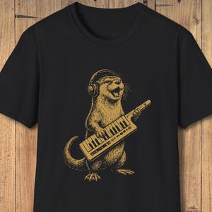 May include: Black t-shirt with a cartoon otter illustration. The otter, in tan, wears headphones and plays a keyboard. The keyboard has the text "OTTER OF WOOD". The design is in a detailed illustration style.