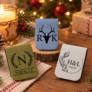 May include: Three rectangular magnetic clips with textured surfaces. The blue clip features a deer head silhouette with the letters R and K. The olive green clip displays the letter N, a laurel wreath, and the text NIRACLE. The white clip shows floral art, H&L, and 12.20.2025.