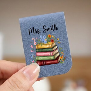 Op de afbeelding: Een blauwe magnetische boekenlegger met een getextureerd oppervlak met een stapel van vijf kleurrijke boeken bekroond met kleine bloemen en de tekst Mrs. Smith in zwarte schrijfletters boven de illustratie.