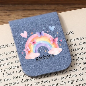 Puede incluir: Un marcapáginas magnético azul con un diseño de arcoíris, mariposas y corazones. El marcapáginas tiene el nombre "Barbara" impreso. El marcapáginas está colocado en un libro abierto.
