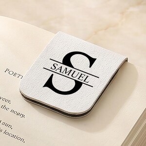以下が含まれることがあります： 「SAMUEL」の名前と「S」の文字が入った白黒のパーソナライズされたブックマークが、開いた本の上に置かれています。コーヒーカップが近くにあります。ブックマークは明るい色の素材で作られています。