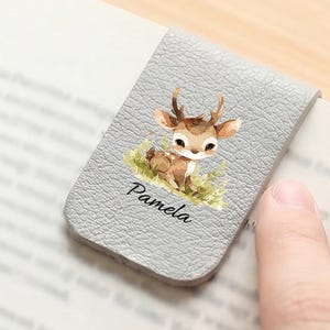 May include: A gray leather bookmark with a watercolor illustration of a fawn on green grass. The name "Pamela" is printed below the fawn. The bookmark is placed in an open book.