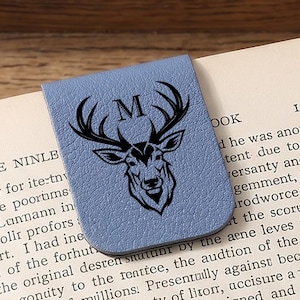 May include: A pale blue leather bookmark featuring a black stag's head and the letter "M". The bookmark is on the pages of a book, offering a stylish way to mark a page. A classic gift.
