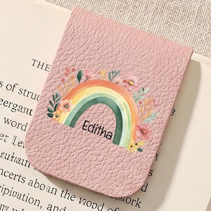 May include: A pink leather bookmark with a rainbow design and the name "Editha" printed on it. The bookmark is placed on an open book.