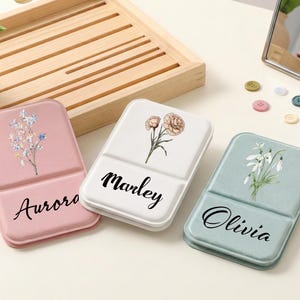 Op de afbeelding: Drie rechthoekige opvouwbare etuis in roze, wit en saliegroen. Elk etui is voorzien van een bloemenillustratie en een gepersonaliseerde naam in zwart schrift: Aurora op het roze etui, Marley op het witte en Olivia op het saliegroene etui.