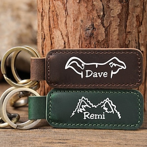May include: Two leather keyrings with metal rings. One is dark brown with a white dog silhouette and the name "Dave." The other is green with a mountain range design and the name "Remi."