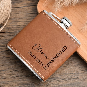 May include: A brown leather flask with a silver cap and trim. The flask is personalized with the name "Oliver", the date "20.10.2025", and the word "Groomsmen" in black lettering. The flask is on a wooden surface.