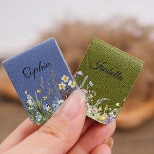 May include: Two rectangular magnetic bookmarks with a textured finish. One is blue with the name Sophia and a floral print, and the other is olive green with the name Isabella and a floral print featuring small yellow, white, and purple flowers.