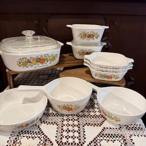 May include: A collection of vintage white ceramic cookware with a colorful fruit and vegetable design. Includes a casserole dish with a glass lid, several rectangular baking dishes, and handled bowls. The text "La Madeleine" is visible.