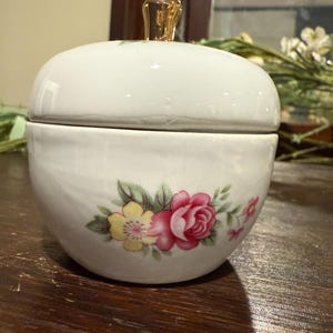 May include: A white ceramic trinket box with a lid, featuring a gold-colored knob. The box is adorned with a floral design in shades of pink, yellow, and green. The box is round and has a glossy finish.