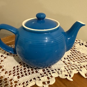 May include: A vibrant blue ceramic teapot with a matching lid and handle. The teapot has a white rim and spout. It sits on a white lace doily on a wooden surface. A classic kitchenware item.