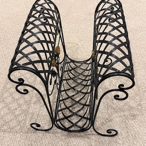 May include: Black metal magazine rack with a decorative lattice design. The rack has two curved compartments and ornate scrollwork feet. Small gold-colored accents are visible on the front.