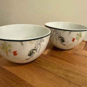 May include: Two white ceramic bowls with a black rim and floral and rooster design. The bowls are on a wooden surface. The bowls have a light blue and green floral design with red rooster accents.