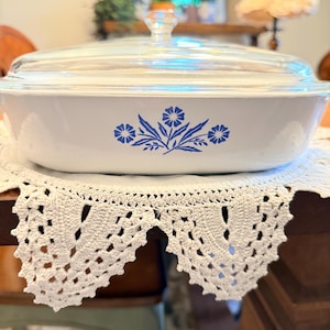 May include: A white casserole dish with a clear glass lid. The dish features a blue floral design on the front. The dish is sitting on a white crocheted doily on a wooden table. A vintage kitchenware item.