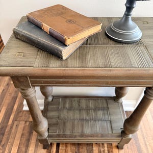 May include: A wooden side table with a woven top and lower shelf, holding two antique books. The books are brown and gray, with visible wear. A decorative lamp with a black base and patterned shade sits on the table.