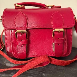 May include: A red leather satchel with a top handle and a long, adjustable shoulder strap. The bag features two front buckle closures and gold-colored hardware. The bag is approximately 8 inches wide and 6 inches tall.