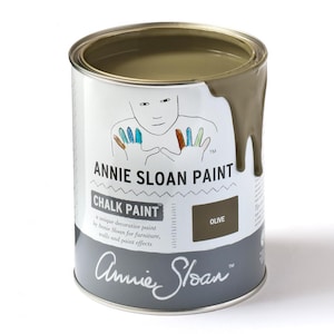 Puede incluir: Una lata de pintura a la tiza Annie Sloan en color Oliva. La lata plateada tiene una etiqueta blanca con un dibujo de una persona con huellas de manos de colores. La pintura es verde oliva y ha goteado por el lateral.