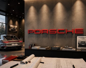 Op de afbeelding: Een werkruimte met architecturale blauwdrukken, pennen en een rood notitieboek op een bureau. Op de achtergrond toont een betonnen muur een groot rood PORSCHE-bord boven een plank met boeken en een modelauto, nabij een zilveren sportwagen.