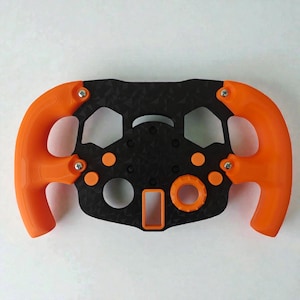 Könnte beinhalten: Ein schwarz-oranges Lenkrad mit geometrischem Design. Das Lenkrad hat orangefarbene Griffe und Akzente, mit einer schwarzen Mitte mit strukturiertem Muster und verschiedenen kreisförmigen und rechteckigen Öffnungen. Befestigt mit silbernen Schrauben.