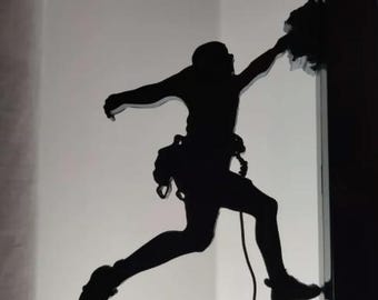 May include: A black silhouette of a rock climber in action, mid-climb. The figure is attached to a rope and gear, with one arm reaching up and the other extended. The background is a gradient of white and gray.