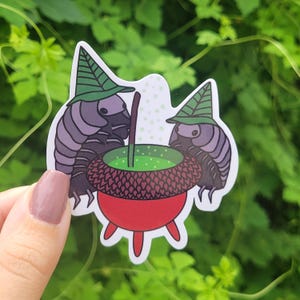May include: A sticker featuring two cartoon pill bugs wearing green witch hats, gathered around a red cauldron filled with green liquid. The pill bugs are purple and gray, and the cauldron has a brown acorn-shaped top.