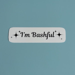 Puede incluir: Una pegatina blanca con esquinas redondeadas y el texto "I'm Bashful" en negro, con una fuente cursiva. Diseños de estrellas negras flanquean el texto. La pegatina está sobre un fondo azul claro.