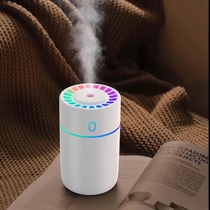 Op de afbeelding: Een witte, cilindrische etherische olie diffuser met een regenboogkleurige lichtring rond de bovenkant en een blauwe lichtband aan de onderkant. De diffuser geeft een stroom damp af. Het apparaat staat op een tijdschrift.