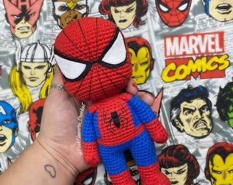 Puede incluir: Un juguete de Spiderman de crochet en rojo y azul, con un diseño de telaraña blanca en el pecho. El juguete está siendo sostenido en una mano contra un fondo de personajes de Marvel Comics.